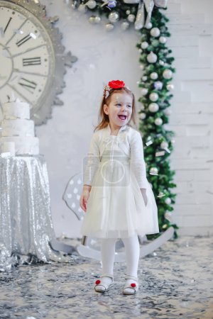 Téléchargez les photos : Jolie petite fille dans une robe blanche avec un bandana rouge sur la tête se tient près du sapin de Noël - en image libre de droit