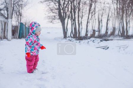 Téléchargez les photos : Petite fille en costume d'hiver lumineux debout sur la rue enneigée en hiver - en image libre de droit