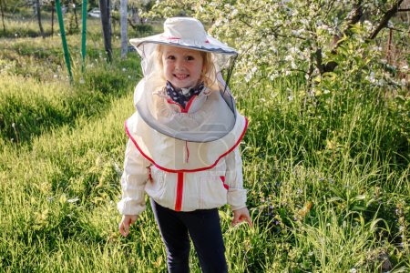 Photo for A little girl in a beekeeper costume helps in an apiary caring for bees - Royalty Free Image