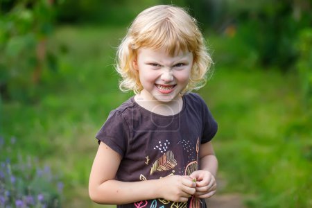 Foto de Niña enojada retorciendo su cara en el parque en el fondo de hierba verde. Retrato de una linda niña con el pelo rubio rizado, jugando en el jardín - Imagen libre de derechos