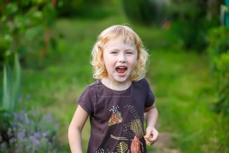 Téléchargez les photos : Petite fille mignonne criant dans le parc avec les yeux plissés. Portrait d'une jolie petite fille dans le jardin par une journée d'été ensoleillée - en image libre de droit