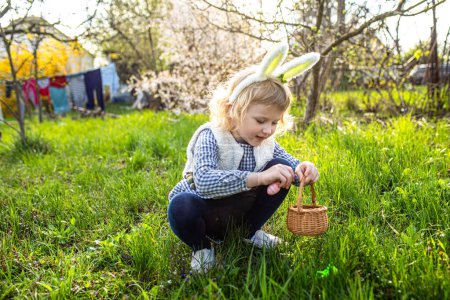 Foto de Niña pequeña con orejas de conejo encontrar y recoger el huevo en la búsqueda de huevos de Pascua en el jardín - Imagen libre de derechos