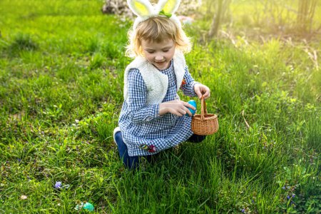 Téléchargez les photos : Fille portant des oreilles de lapin jouant à la chasse aux œufs à Pâques. Un enfant célèbre Pâques. Fille assise sur l'herbe avec un panier plein d'œufs colorés. Chasse aux œufs de Pâques dans le jardin - en image libre de droit