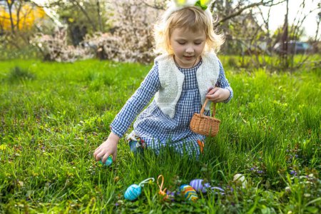 Foto de Caza de huevos de Pascua. Niña con cestas en las manos recogiendo huevos de colores en el parque - Imagen libre de derechos