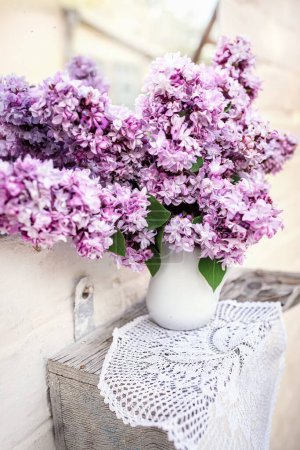 Foto de Ramo de lila púrpura en jarrón de cerámica blanca en servilleta tejida cerca del espejo vintage Decoración para el hogar con flores frescas. - Imagen libre de derechos