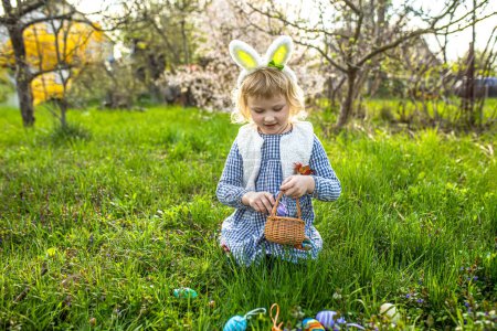 Foto de La alegría de Pascua. niña alegre reúne huevos vibrantes en la cesta en medio de la vegetación del parque. Caza de huevos de Pascua en jardín - Imagen libre de derechos