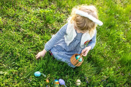 Foto de Niña pequeña con orejas de conejo encontrar y recoger el huevo en la búsqueda del huevo de Pascua en el jardín de la caza del huevo de Pascua en el jardín - Imagen libre de derechos