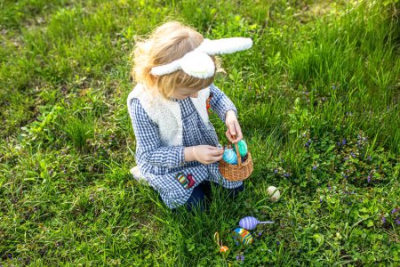 Foto de Tradición pascual. Una chica con una cesta recoge coloridos huevos de Pascua en la hierba. Niño con orejas de conejo. Caza de huevos de Pascua en jardín - Imagen libre de derechos