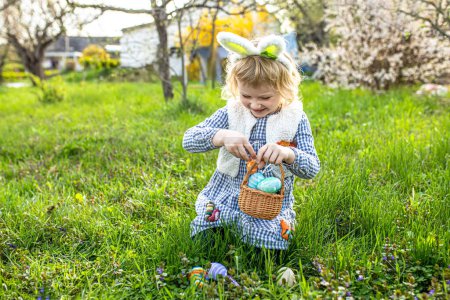 Téléchargez les photos : Traditions pascales ludiques. enfant heureux s'engage dans une chasse aux œufs, remplir le panier avec des symboles de printemps et de renouvellement. Chasse aux œufs de Pâques dans le jardin - en image libre de droit