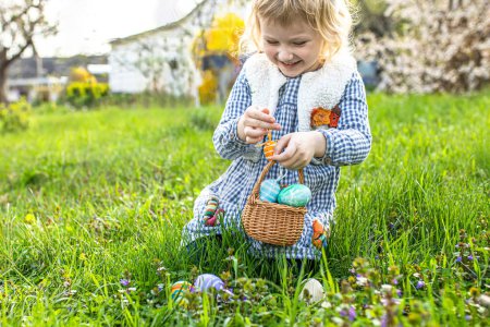 Téléchargez les photos : Petite fille avec un sourire rayonnant cherche des œufs de Pâques, chasse aux œufs de Pâques dans le jardin - en image libre de droit