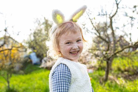 Photo for Little girl smiling happily wearing Bunny ears Easter egg hunt in garden - Royalty Free Image