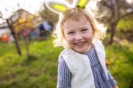 Foto de Retrato de una niña rubia con orejas de conejito sonriendo en el jardín. Caza de huevos. - Imagen libre de derechos