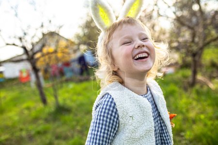 Téléchargez les photos : Portrait de petite fille blonde portant des oreilles de lapin souriant dans le jardin. Chasse aux œufs de Pâques. - en image libre de droit