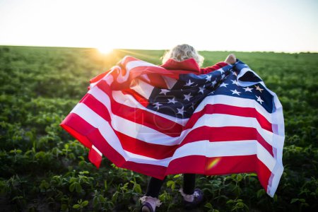 Foto de Bandera de América en hombros de un niño. chica en el fondo de la puesta del sol. Little American celebra a EE.UU. Día de la Independencia el 4 de julio - Imagen libre de derechos