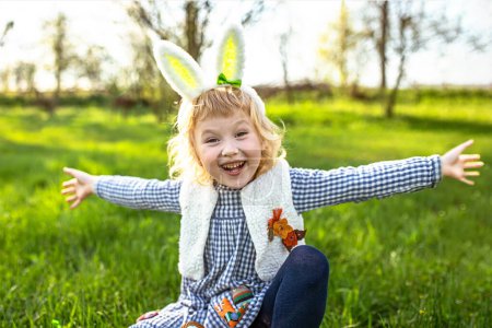 Téléchargez les photos : Une heureuse petite fille enfilait des oreilles de lapin de Pâques et étendait ses bras sur les côtés en se réjouissant lors d'un jeu de récupération d'œufs. Chasse aux œufs de Pâques dans le jardin - en image libre de droit