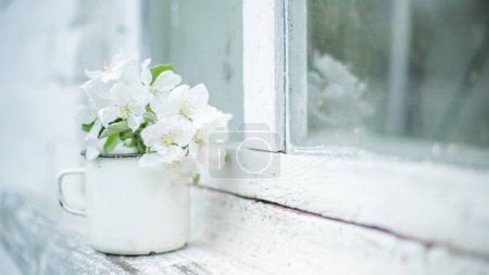 Téléchargez les photos : Nature morte avec tasse avec branche fleurie de pommier sur le rebord d'une fenêtre blanche près d'une vieille fenêtre en bois. Image vintage. Fonds printanier. - en image libre de droit