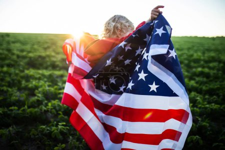 Téléchargez les photos : Fille avec un drapeau américain dans le fond du coucher de soleil. Rayons de soleil sur champ vert de soja. Vacances patriotiques. - en image libre de droit