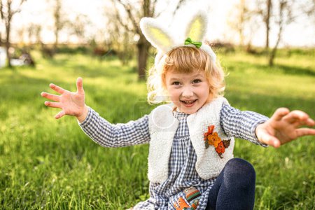 Téléchargez les photos : Joyeux petite fille mis portant des oreilles de lapin de Pâques et étendre ses bras sur les côtés se réjouissant lors d'un jeu de récupération d'œufs. Oeufs de Pâques chasse dans le jardin. - en image libre de droit