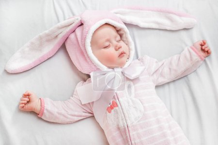 Foto de Niña recién nacida en traje de dormir con orejas de conejito de Pascua descansa durante la siesta con los brazos extendidos a los lados. - Imagen libre de derechos