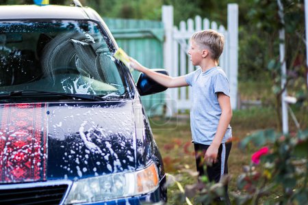 Téléchargez les photos : Un adolescent caucasien lave sa voiture en la savonnant avec du détergent pour lave-auto dans la cour de la maison un jour d'été. Aide à l'enfance - en image libre de droit