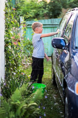 Téléchargez les photos : Un adolescent en survêtement lave un minivan avec de l'eau savonneuse dans la cour d'une maison en été. La notion de responsabilité dans les affaires quotidiennes de la famille - en image libre de droit