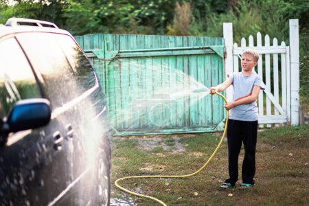Foto de Un adolescente lava un coche vertiendo agua sobre él con agua de una manguera de jardín. El concepto de cooperación familiar - Imagen libre de derechos