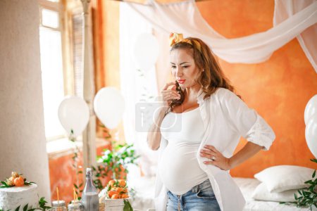 Photo for Young pregnant woman in white shirt celebrating baby blessing in bedroom with orange walls decorated with balloons. Baby shower concept. - Royalty Free Image