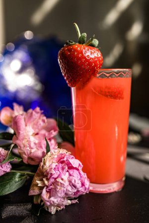 Téléchargez les photos : Jus de fraise dans un verre sur fond flou avec des fleurs. Eau infusée à la fraise et au citron vert, associée à des pivoines pour une hydratation rafraîchissante et saine. Concept de délice nutritif - en image libre de droit