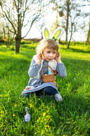 Alegre niña disfruta de la búsqueda del huevo de Pascua, llenando alegremente su cesta con coloridas delicias en medio de las festividades al aire libre.