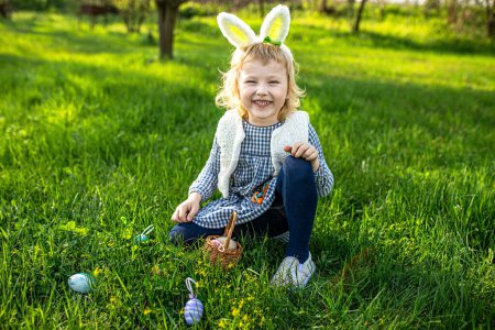 Nettes kleines Mädchen, geschmückt mit Hasenohren, kichert am Ostertag fröhlich in einem blühenden Garten und umarmt das saisonale Fest mit Spaß bei der Eiersuche.