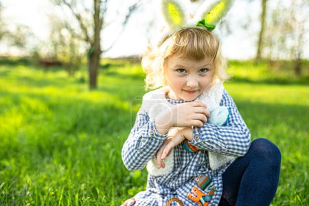Glückliches kleines Mädchen, mit Hasenohren vor der Kulisse eines lebendigen Gartens am Ostertag, das mit Freude an der Eiersuche das saisonale Fest feiert.