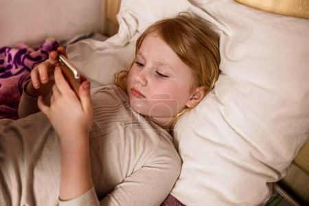 Photo for Cute little girl lying in bed and playing with a mobile phone - Royalty Free Image