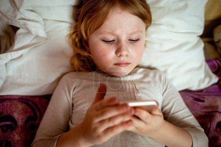 Téléchargez les photos : Petite fille utilisant le téléphone mobile dans le lit à la maison. - en image libre de droit