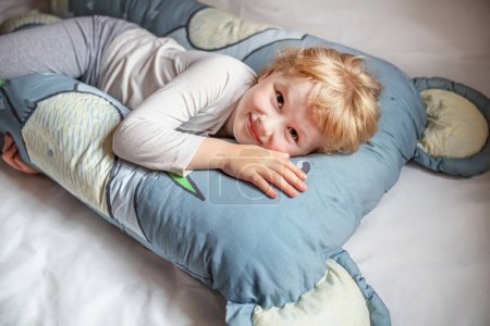 Foto de Alegre niño pequeño con el pelo rubio se encuentra cómodamente en un cojín de gran tamaño en un dormitorio adornado con tonos azules y grises tranquilos. - Imagen libre de derechos