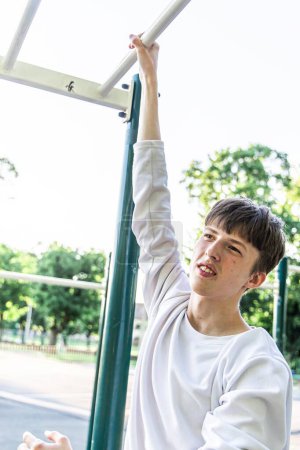Dieses Bild zeigt einen jungen Mann beim Klimmzug an einer Stange auf einem Outdoor-Trainingsplatz in einem Park. Er trägt ein weißes langärmeliges Hemd, die Bar ist grün-weiß. Im Hintergrund ist Laub.