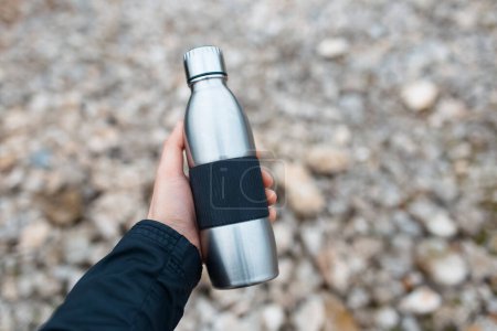 Foto de Close-up of male hand holding steel thermo water bottle on background of blurred rocky ground. - Imagen libre de derechos