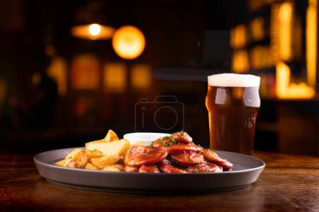Foto de Porción de embutidos con papas fritas rústicas y salsa barbacoa y pinta de curry de cerveza pálida en fondo borroso pub - Imagen libre de derechos