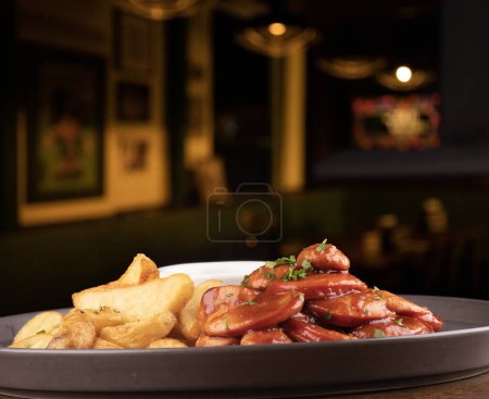 Foto de Porción de embutidos con papas fritas rústicas y salsa barbacoa y curry con pub borroso en el fondo - Imagen libre de derechos