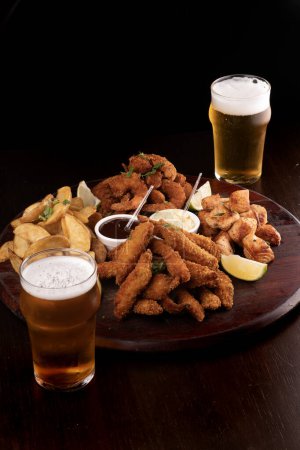 portion of seafood with breaded fish and shrimp and grilled salmon with french fries and two glasses of craft beer dark background portrait