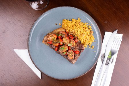 Foto de Dos trozos de solomillo cocido con ensalada de tomate cherry y farofa en una cena en el asador vista superior con cubiertos - Imagen libre de derechos