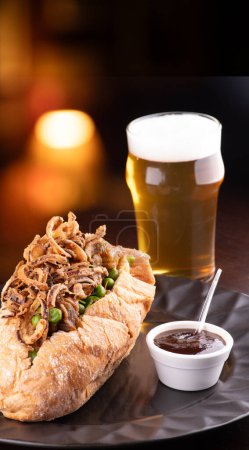 beef stew sandwich with crispy onions and vegetables and bbq sauce with half pint of beer and blurred background