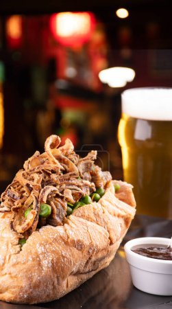 beef stew sandwich with crispy onions and vegetables and barbecue sauce with half a pint of beer and blurred background