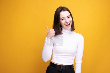 Foto de Feliz chica adulta mostrando los pulgares arriba con una camiseta blanca sobre un fondo amarillo. - Imagen libre de derechos
