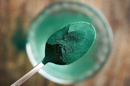 Green spirulina powder on a spoon above a glass of water