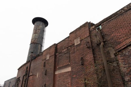 Exterior brick wall of an industrial facility with deeply weathered smokestack, gray sky winter, horizontal aspect