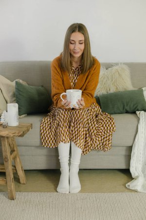 Téléchargez les photos : Femme souriante positive buvant tasse de café aromatique assis sur le canapé à la maison. Confort ambiance chaleureuse - en image libre de droit