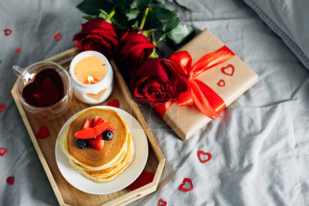 Valentine's day breakfast concept. Pancakes with berries, roses flowers, cup of tea, gift box and candle in candlestick. Top view.