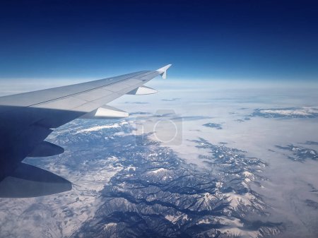 Téléchargez les photos : Plane flight above the Carpathian Mountains snowy peaks. Blue skyline and airplane wing seen through the window - en image libre de droit