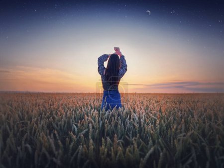 Téléchargez les photos : Fille dans le champ de blé doré au coucher du soleil. Beau paysage crépusculaire sous le ciel étoilé d'été avec croissant de lune. Scène naturelle magique, concept de liberté - en image libre de droit