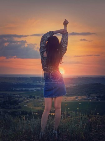 Foto de Concepto de juventud salvaje y libre con una chica relajada, la vista trasera se levanta con las manos en el viento frente a la hermosa puesta de sol de verano en la cima de una colina que mira sobre el valle - Imagen libre de derechos